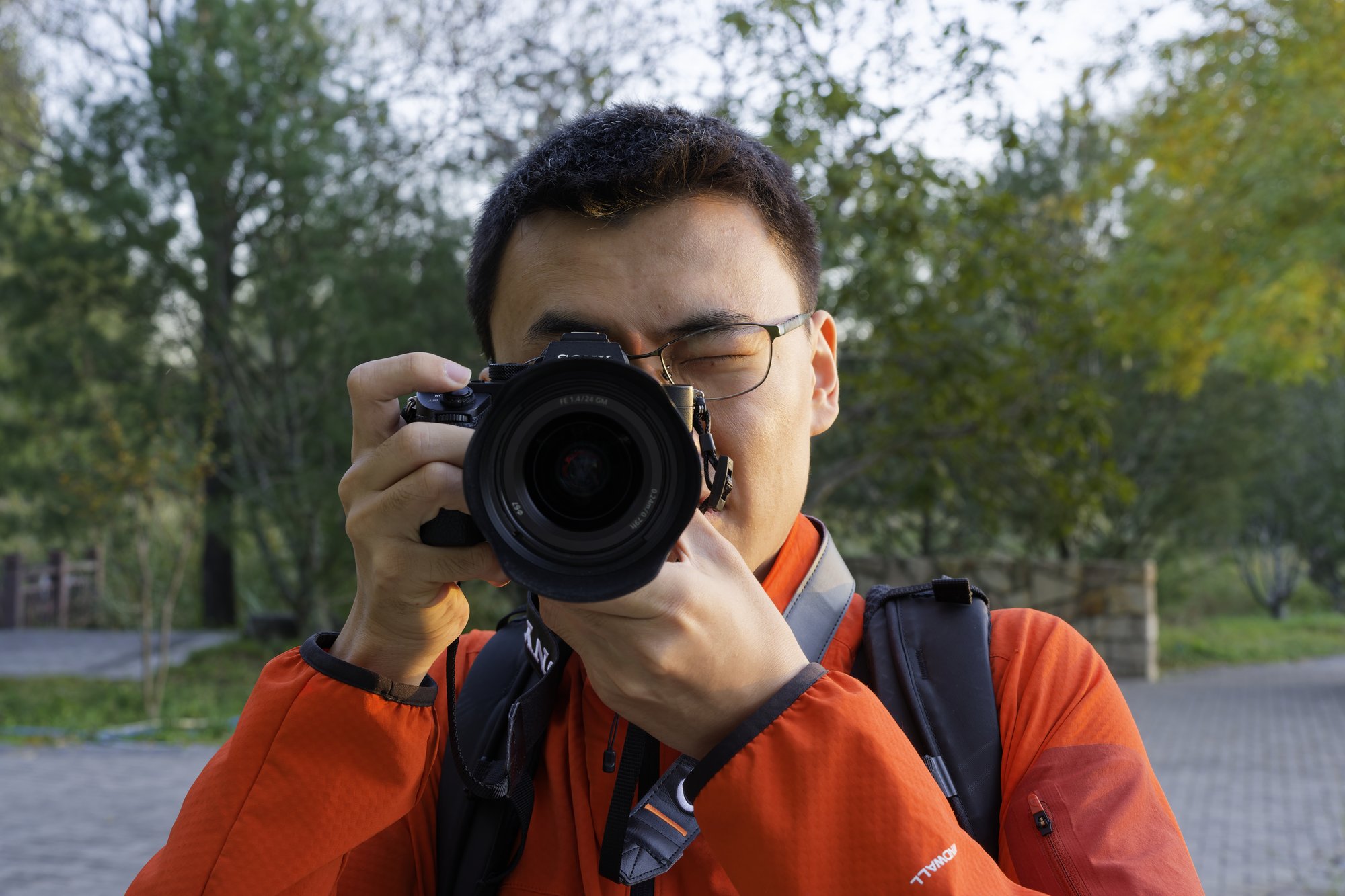 Wang Yinbo, in glasses and an orange jacket, holds a Sony camera with a wide lens up to one eye, with trees and a paved path blurred behind in Beiwu Park.