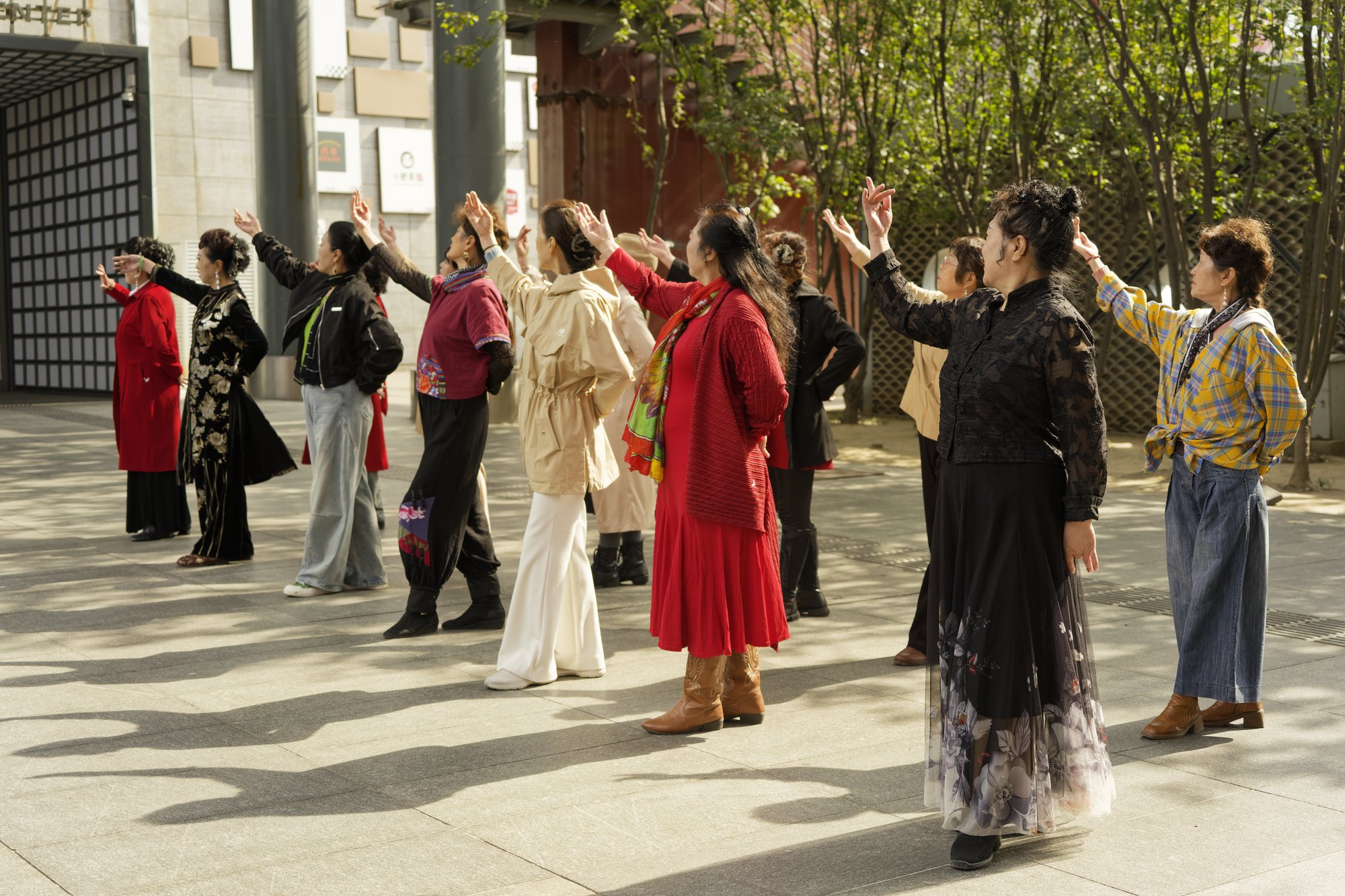 A dozen women in red dresses, flowered skirts, trench coats and plaid shirts stand in loose rows on a sunny paved plaza, each raising one arm overhead in the same dance move, with trees and a grey building behind.