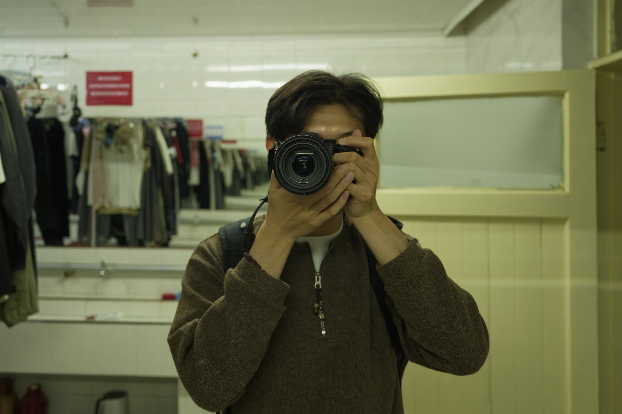 The photographer, in a brown zip-neck sweater with a backpack strap over one shoulder, holds a Sony camera up to the face in front of a mirror, the lens hiding the eyes; tiled walls, sinks and racks of hanging clothes stretch away behind.
