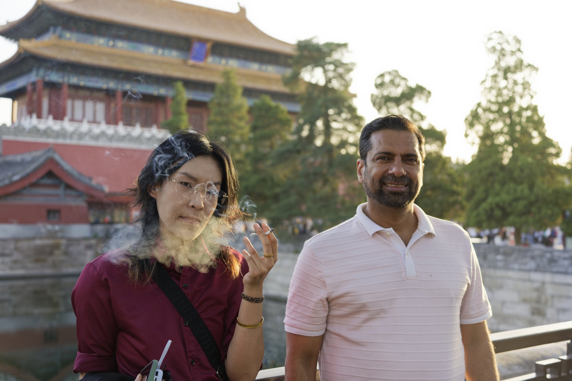 A woman in glasses and a burgundy shirt breathes out a cloud of smoke, a cigarette between her fingers, beside a smiling bearded man in a pale pink polo shirt; they stand at a railing by the moat, with a palace gate tower, red walls and tall pines behind them in warm evening light.