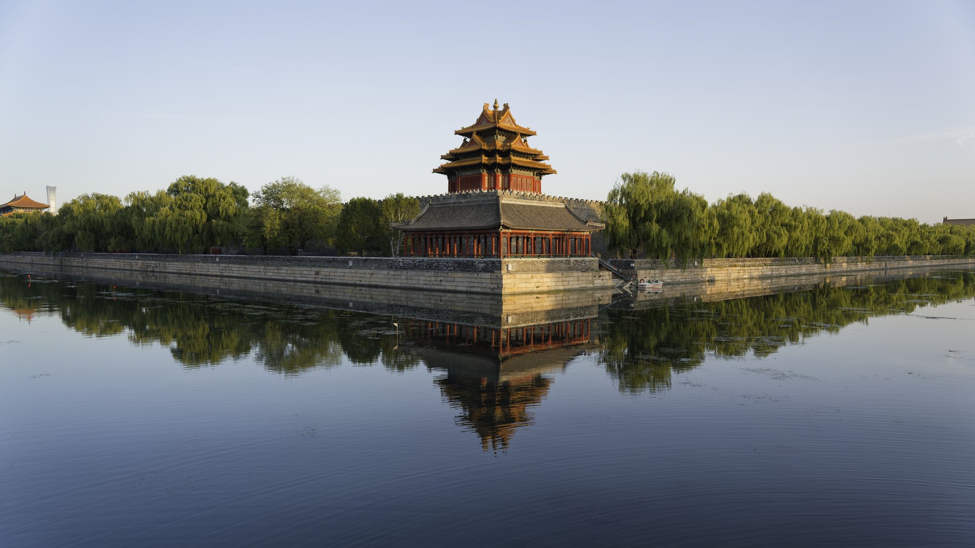 The many-eaved corner tower of the Forbidden City, golden roofs over red walls, rises from the corner of a long stone wall and is mirrored almost perfectly in the calm blue moat, with willows lining both banks under a clear pale sky.