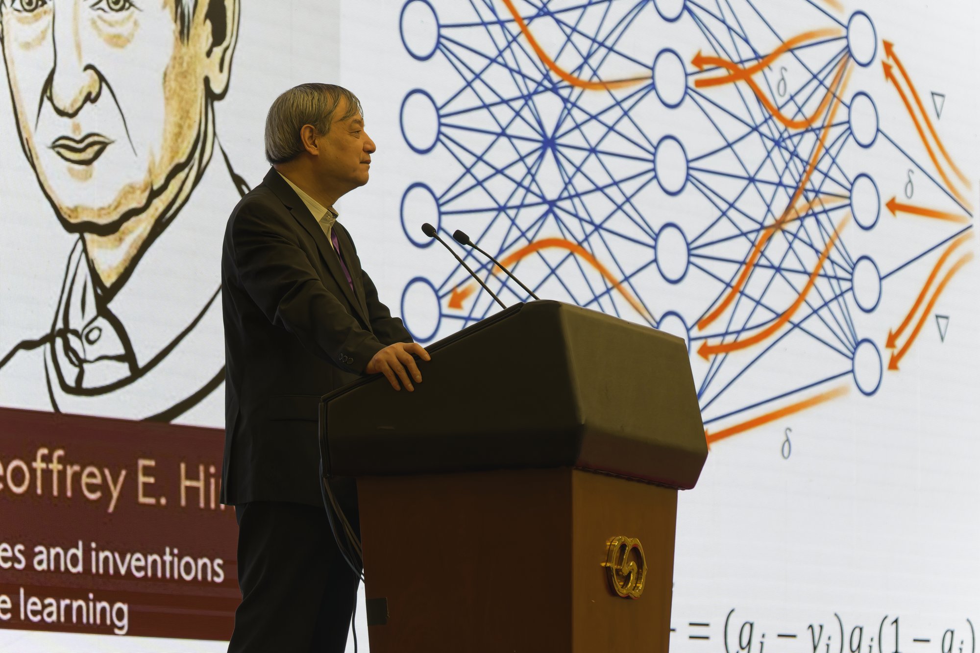 Professor Sun Maosong, grey-haired in a dark suit, stands at a wooden lectern with two microphones and looks to the right; behind him a giant screen shows a sketched portrait of Geoffrey E. Hinton and a neural-network diagram with orange arrows running backwards through it.