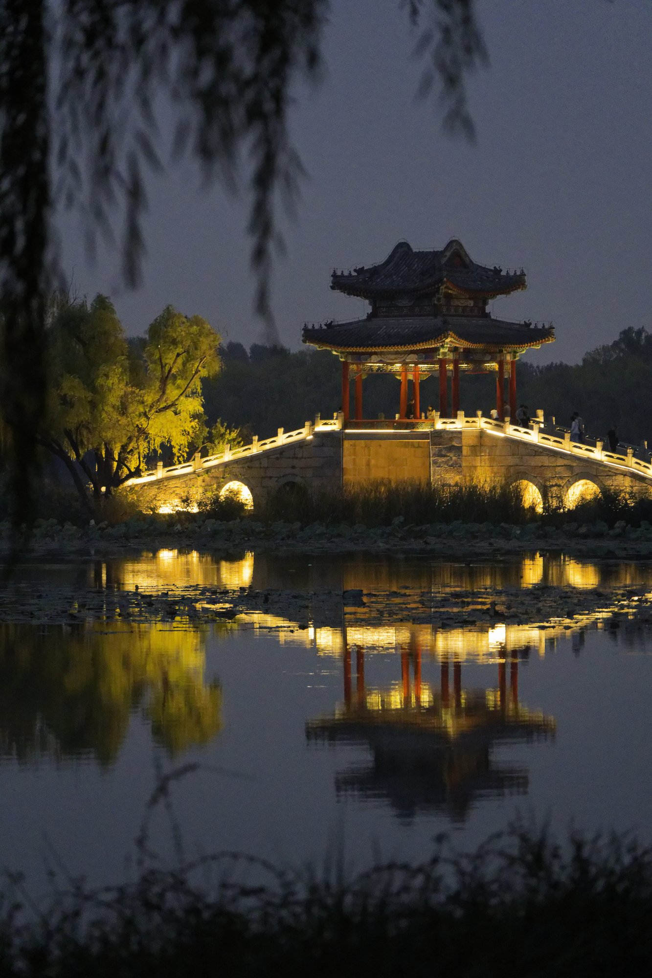 A lit stone bridge with a red-pillared, double-eaved pavilion reflected in a lake at night in the Summer Palace, framed by willow branches.
