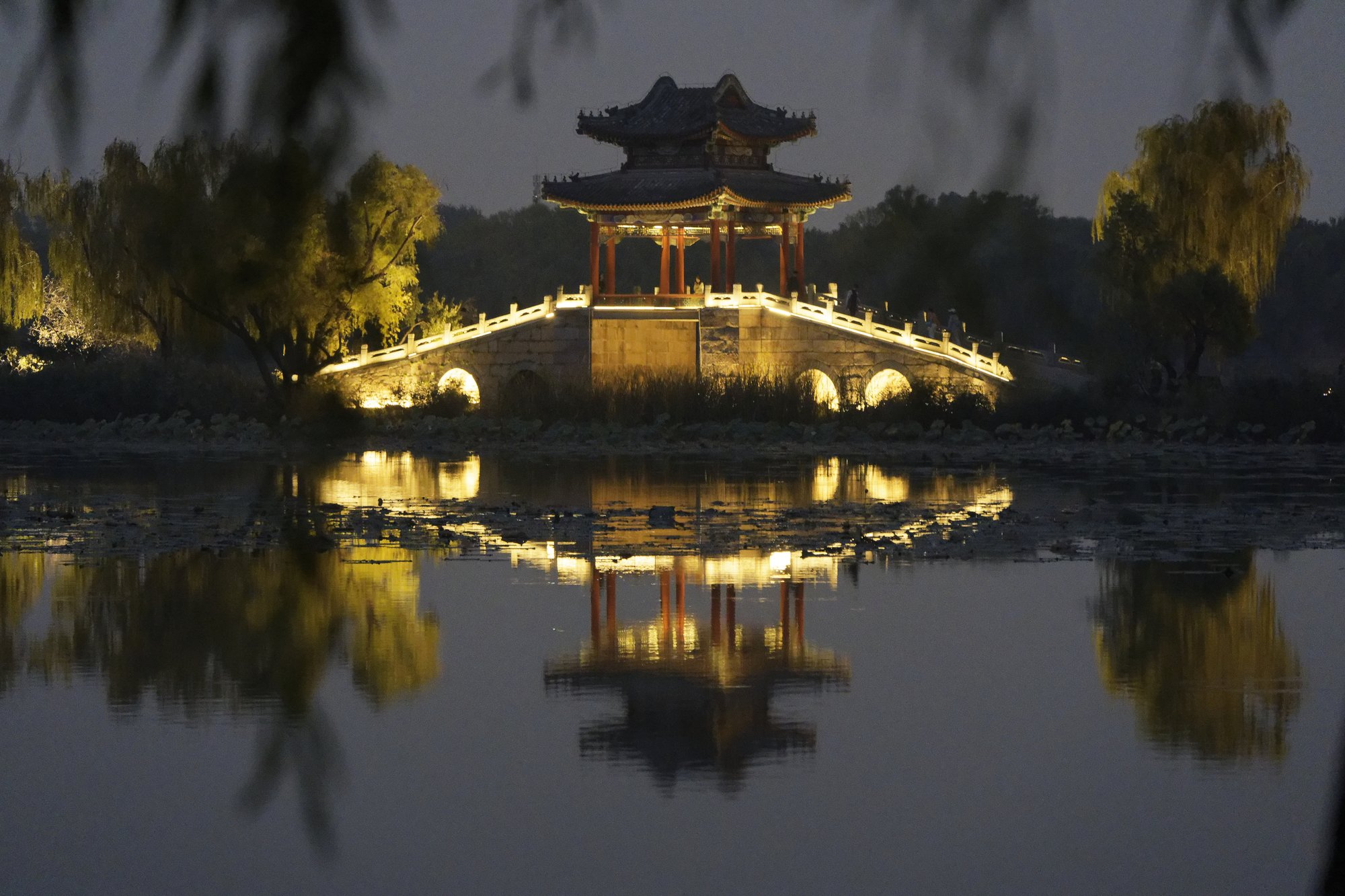 A lit pavilion bridge and illuminated willows reflected in a calm lake at night in the Summer Palace, with blurred willow branches in the foreground.